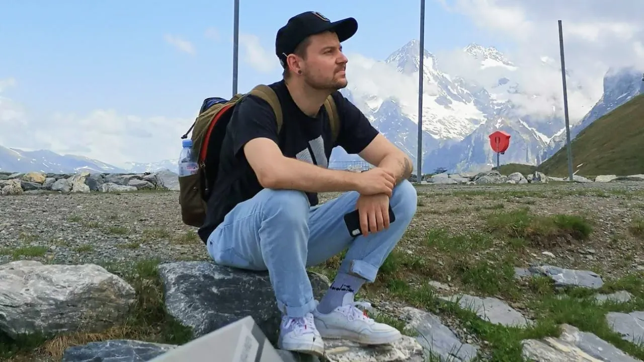 Martin in a black cap and backpack sitting on a rock with snow-covered mountains in the background.